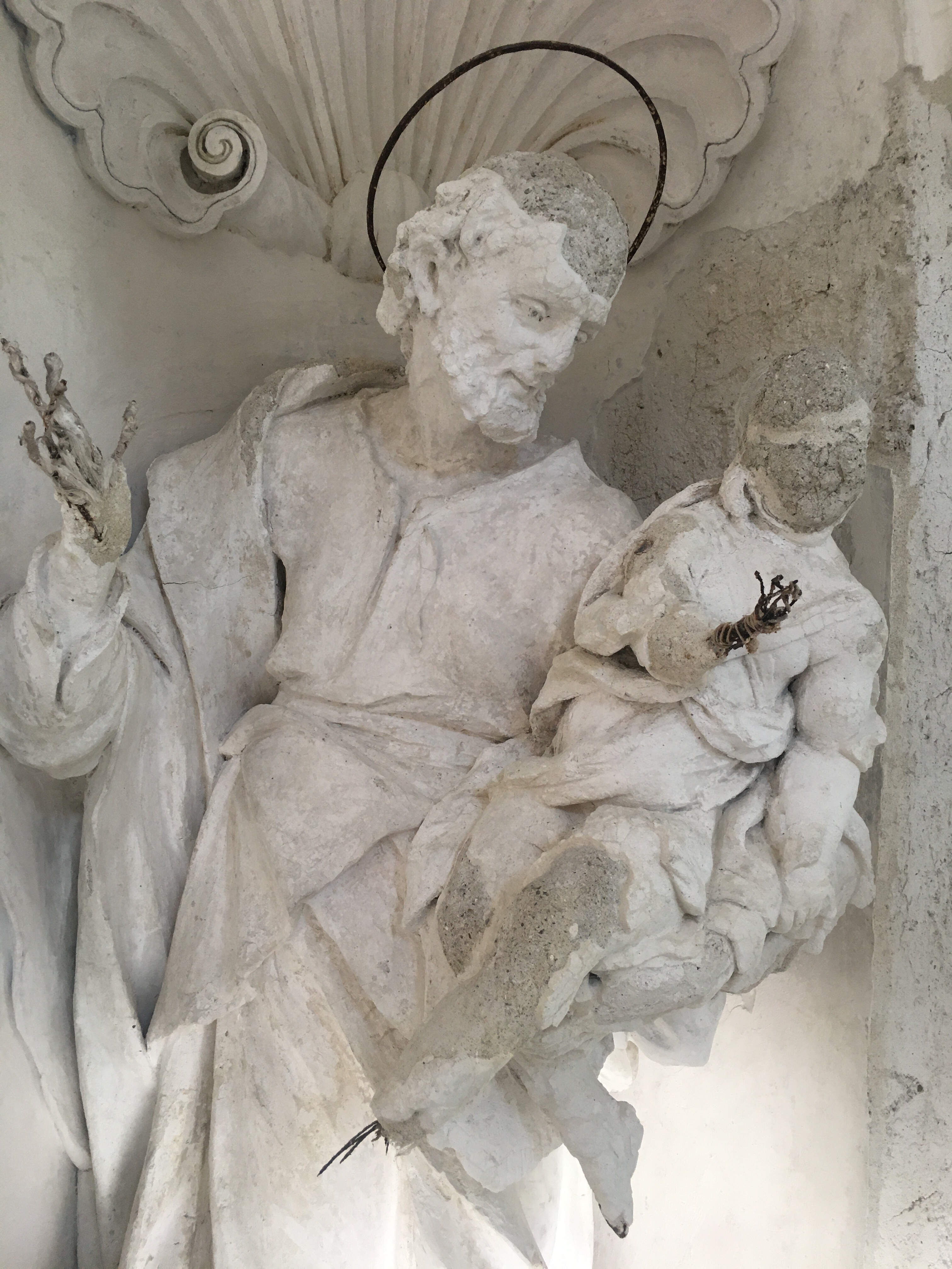 A weathered plaster statue of a bearded saint with a metal halo, holding a child and set in a niche with a scalloped shell design. The statue’s deteriorated condition exposes internal iron bars as well as stucco and white finishing layers.