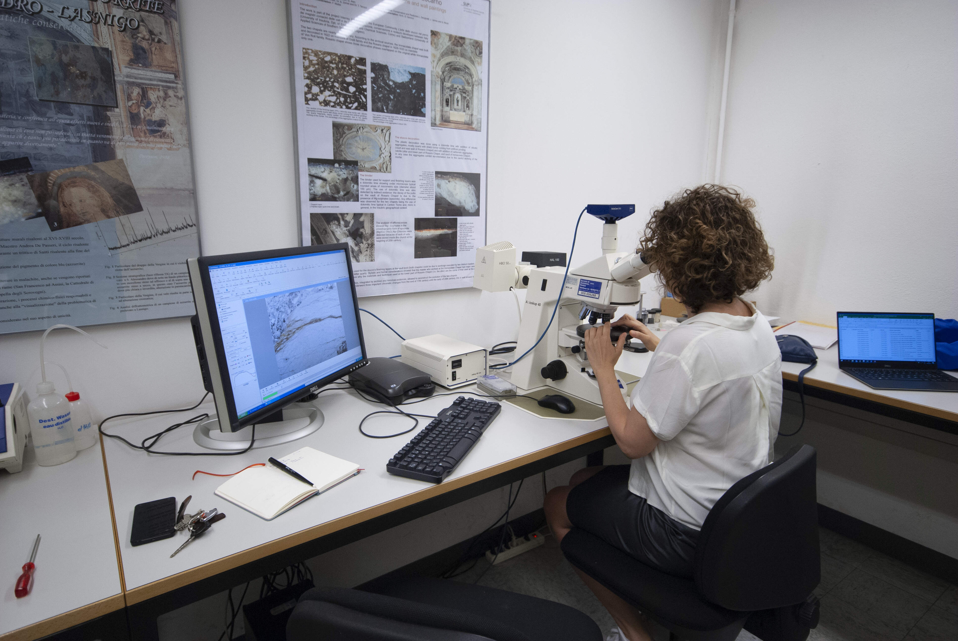 A researcher in a laboratory examines a sample under a microscope. A desktop computer displays a magnified image of the sample.