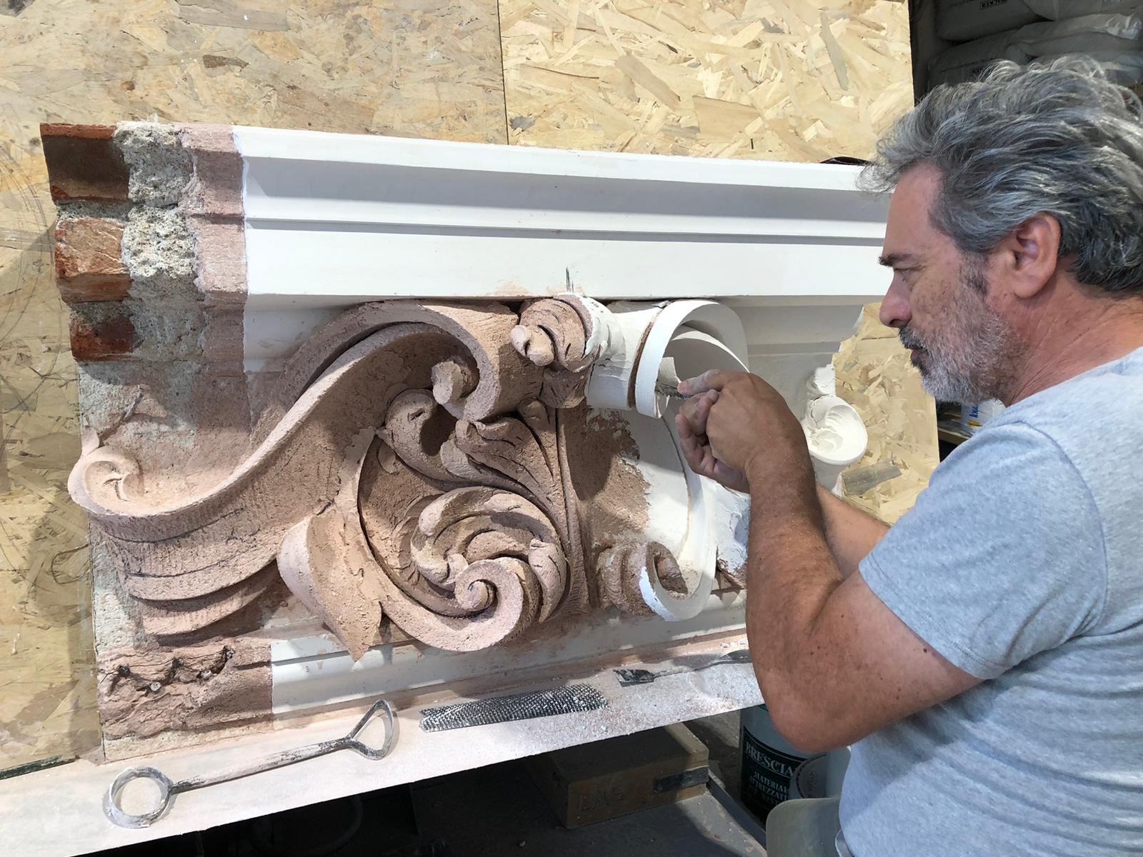 A member of the research team making a stucco decoration replica. A brick support and three layers (lime-gypsum, pozzolana-lime, and a finishing layer of lime and marble powder) are visible, with working tools lying at the base of the decoration.